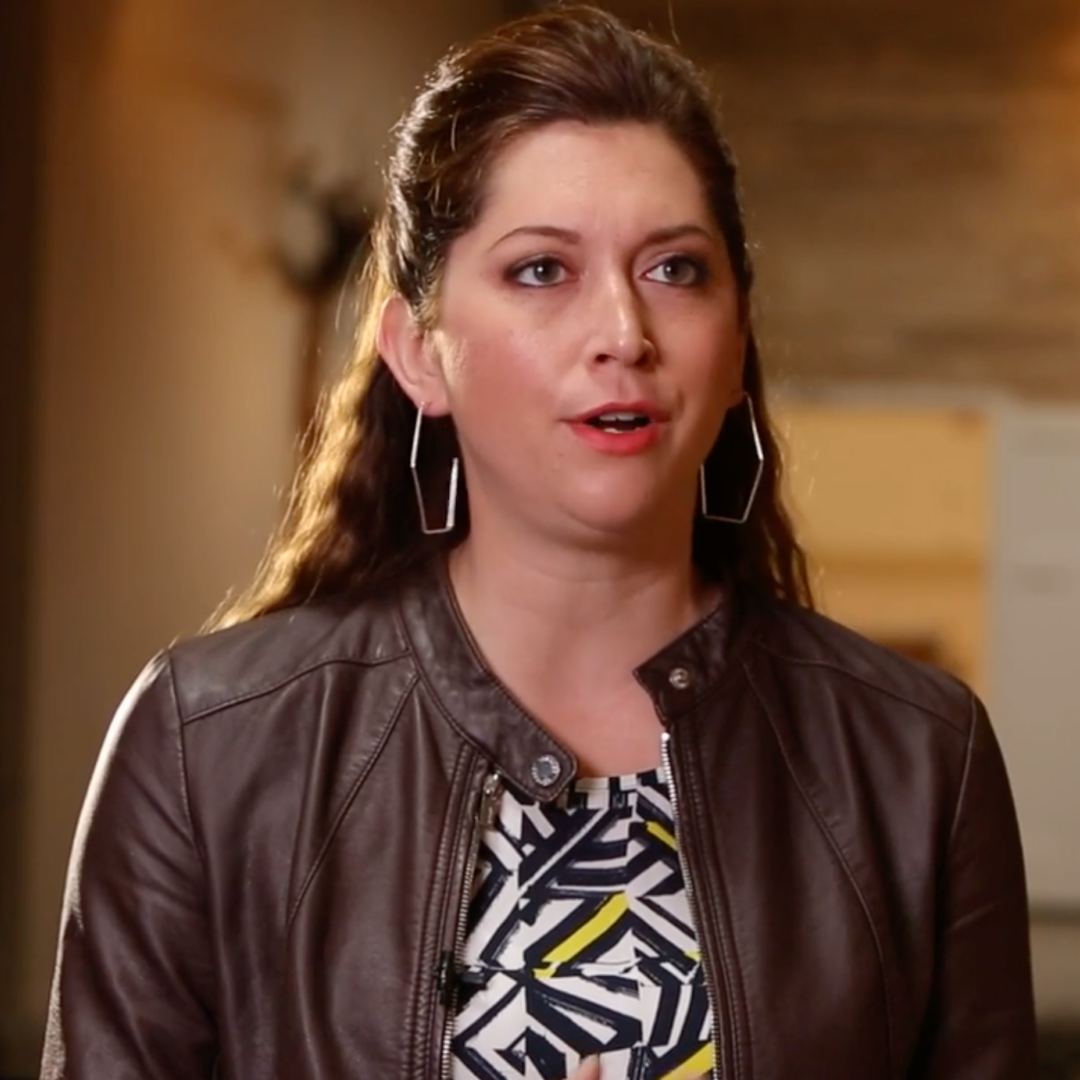 A woman with long brown hair, wearing a brown leather jacket, geometric earrings, and a patterned top, stands indoors speaking with a neutral background behind her.